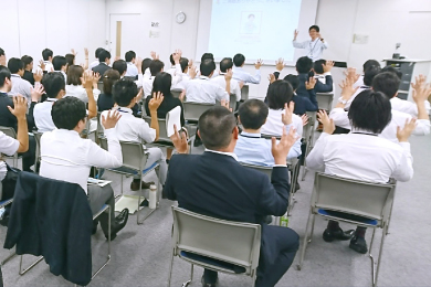 広い会議室で40数名の社員が、障がい理解研修を受講している全体のショット。講師に合わせて、参加者全員が手話に取り組んでいる様子。