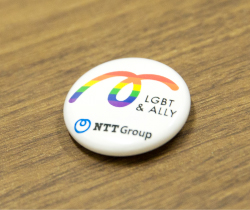NTTグループオリジナルのLGBTアンドALLYアライのバッヂ。バッヂには、LGBTアンドALLYアライとNTTグループの文字と一緒に虹が描かれている。