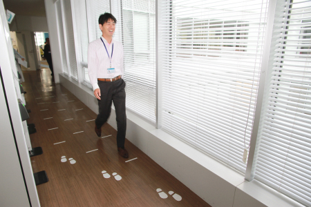 社員が、休憩スペースの床に描かれた印の上をウォーキングしているショット。３種類の中から、自分の身長に合わせた歩幅の印を選んで歩くと程よい運動になります。