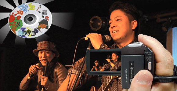 写真：左上の放射状の光りに照らされたDVDと、ボーカルの圭太さんと大平さんがマイクを持ってライブ中にトークをしている様子を手前からビデオカメラで撮影しているところをを合成している。