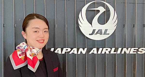 写真：JAPAN AIRLINESのロゴマークをバックに受付で制服を着た女性社員