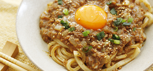 写真：お皿にできあがったキーマカレーうどんが盛られている。トッピングに白ごま、青ネギ、そして中央には卵の黄身が輝いている！