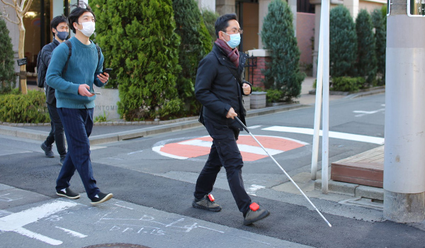 写真：実証実験で狭い道を視覚障がい者と一緒に歩く千野さん