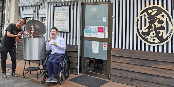 写真：お店の前で店主の毛塚さんと車いすユーザの編集部員が鍋のディスプレイでポーズを取っている