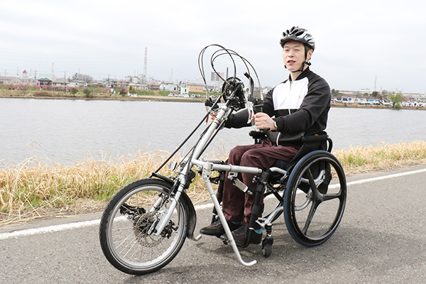 写真：河川敷の道路でアダプタ式のハンドサイクルを自分の車いすに取り付けして走行している編集部員