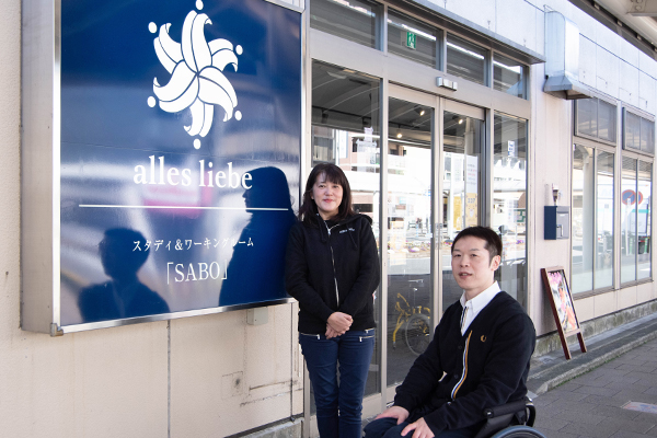 写真：事務所前でユリをモチーフにしたロゴの看板前で記念撮影する青木さんと車いすユーザの編集部員