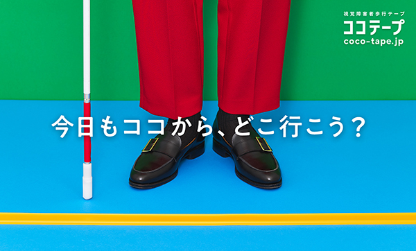 写真：青い床にココテープが横一線に貼られており、その手前に白杖を持って立っている赤いズボンの人の膝から下が映っている。写真にかぶせて全体のセンターに「今日もココから、どこ行こう？」と書いてある