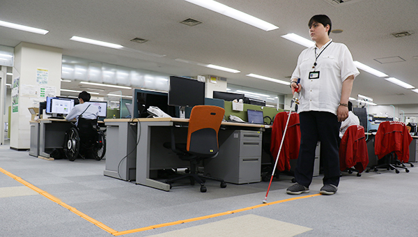 写真：ココテープに白杖を添わせて歩く社員