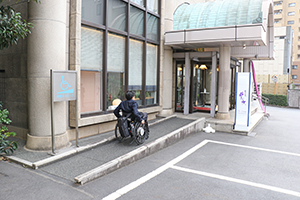 写真：入口ドアに続くスロープをのぼる車いすユーザ
